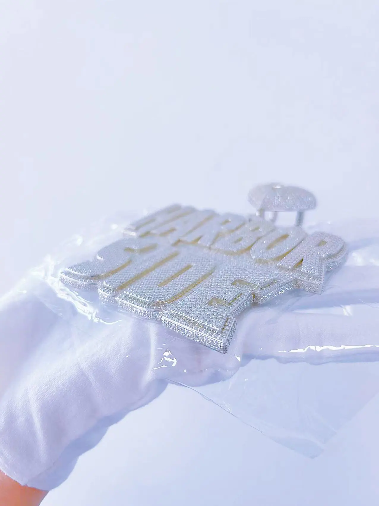 Side view of custom large letter name pendant in 925 silver with VVS moissanite, plated in 18K white gold, held in hand inside clear bag, showing two rows of pavé stones on the side.